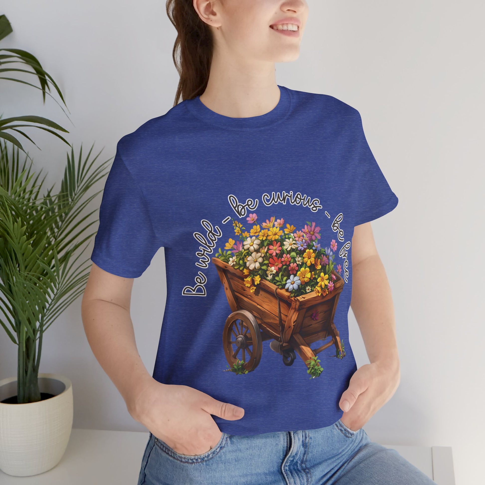 Colorful illustration of a wooden wheelbarrow filled with vibrant flowers, surrounded by the handwritten text 'Be wild - be curious - be free.' This design is intended for t-shirts in various colors, including Aqua, Athletic Heather, Black, Dark Grey Heather, Heather Kelly, Heather Red, Heather True Royal, Natural, Navy, Pink, White, Heather Aqua, Heather Ice Blue, Heather Mauve, and Charity Pink.