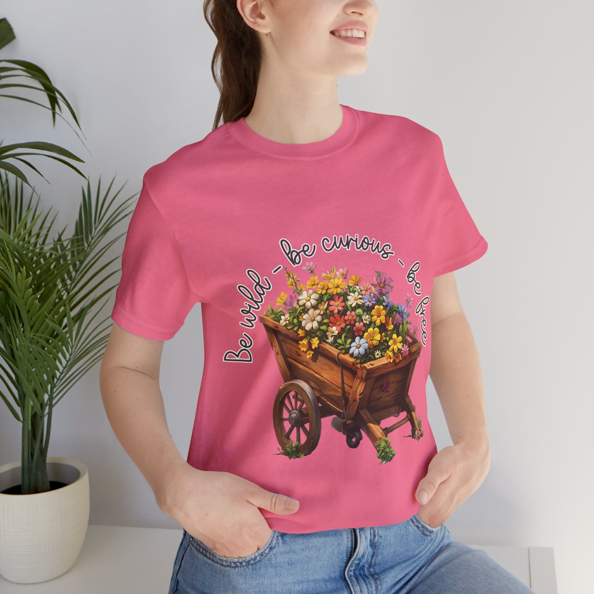 Colorful illustration of a wooden wheelbarrow filled with vibrant flowers, surrounded by the handwritten text 'Be wild - be curious - be free.' This design is intended for t-shirts in various colors, including Aqua, Athletic Heather, Black, Dark Grey Heather, Heather Kelly, Heather Red, Heather True Royal, Natural, Navy, Pink, White, Heather Aqua, Heather Ice Blue, Heather Mauve, and Charity Pink.