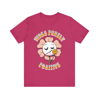 Cute pink flower with a smiling face and a yellow flower in its mouth, accompanied by the text "Vibes Purely Positive." This cheerful design is perfect for t-shirts available in colors Aqua, Berry, Black, Dark Gray, Heather Kelly, Heather Team Purple, Heather True Royal, Navy, White, and Charity Pink.