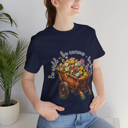 Colorful illustration of a wooden wheelbarrow filled with vibrant flowers, surrounded by the handwritten text 'Be wild - be curious - be free.' This design is intended for t-shirts in various colors, including Aqua, Athletic Heather, Black, Dark Grey Heather, Heather Kelly, Heather Red, Heather True Royal, Natural, Navy, Pink, White, Heather Aqua, Heather Ice Blue, Heather Mauve, and Charity Pink.