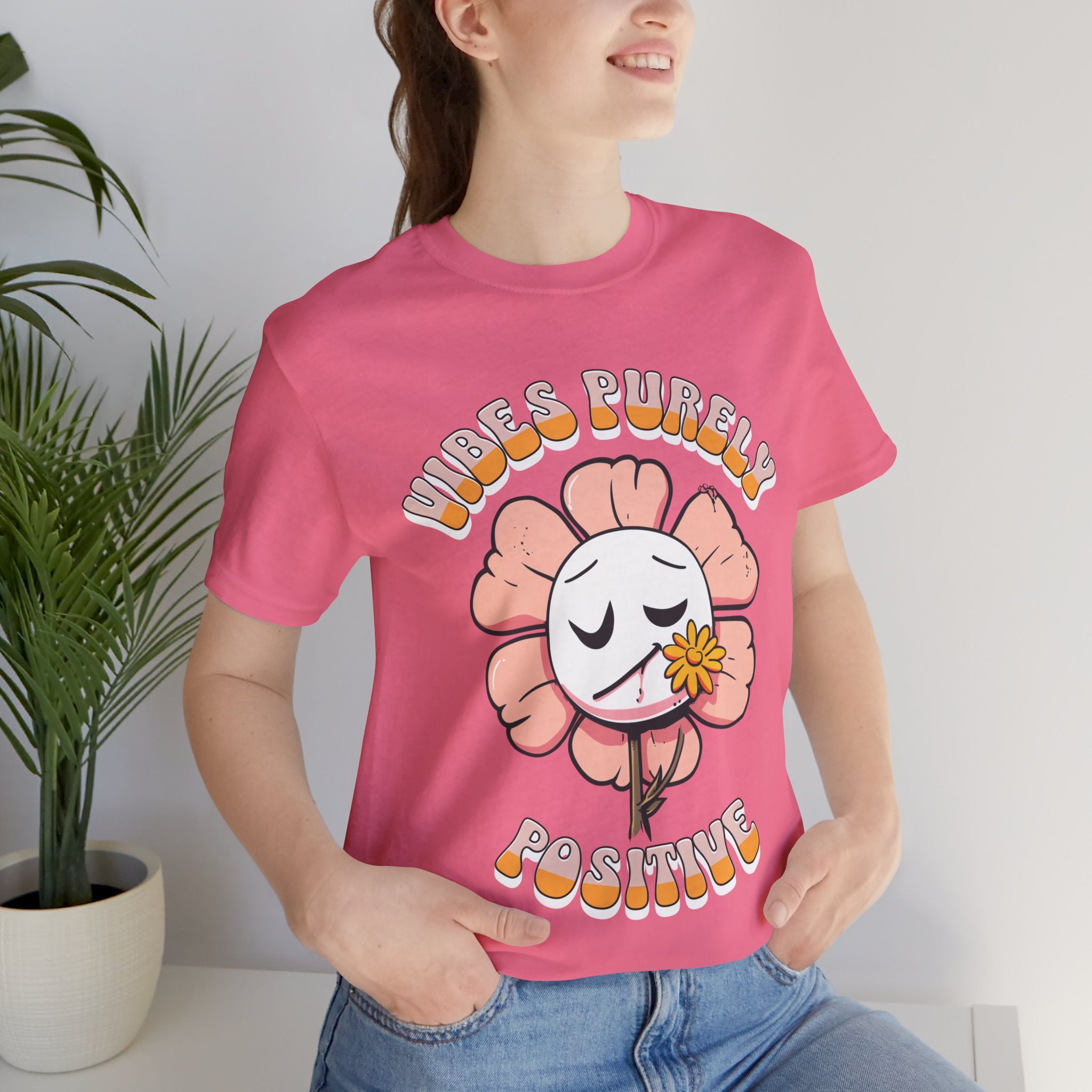 Cute pink flower with a smiling face and a yellow flower in its mouth, accompanied by the text "Vibes Purely Positive." This cheerful design is perfect for t-shirts available in colors Aqua, Berry, Black, Dark Gray, Heather Kelly, Heather Team Purple, Heather True Royal, Navy, White, and Charity Pink.