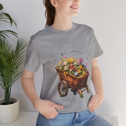 Colorful illustration of a wooden wheelbarrow filled with vibrant flowers, surrounded by the handwritten text 'Be wild - be curious - be free.' This design is intended for t-shirts in various colors, including Aqua, Athletic Heather, Black, Dark Grey Heather, Heather Kelly, Heather Red, Heather True Royal, Natural, Navy, Pink, White, Heather Aqua, Heather Ice Blue, Heather Mauve, and Charity Pink.