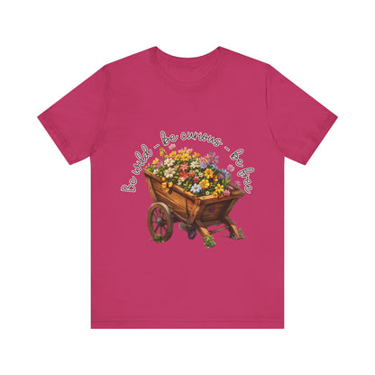 Colorful illustration of a wooden wheelbarrow filled with vibrant flowers, surrounded by the handwritten text 'Be wild - be curious - be free.' This design is intended for t-shirts in various colors, including Aqua, Athletic Heather, Black, Dark Grey Heather, Heather Kelly, Heather Red, Heather True Royal, Natural, Navy, Pink, White, Heather Aqua, Heather Ice Blue, Heather Mauve, and Charity Pink.