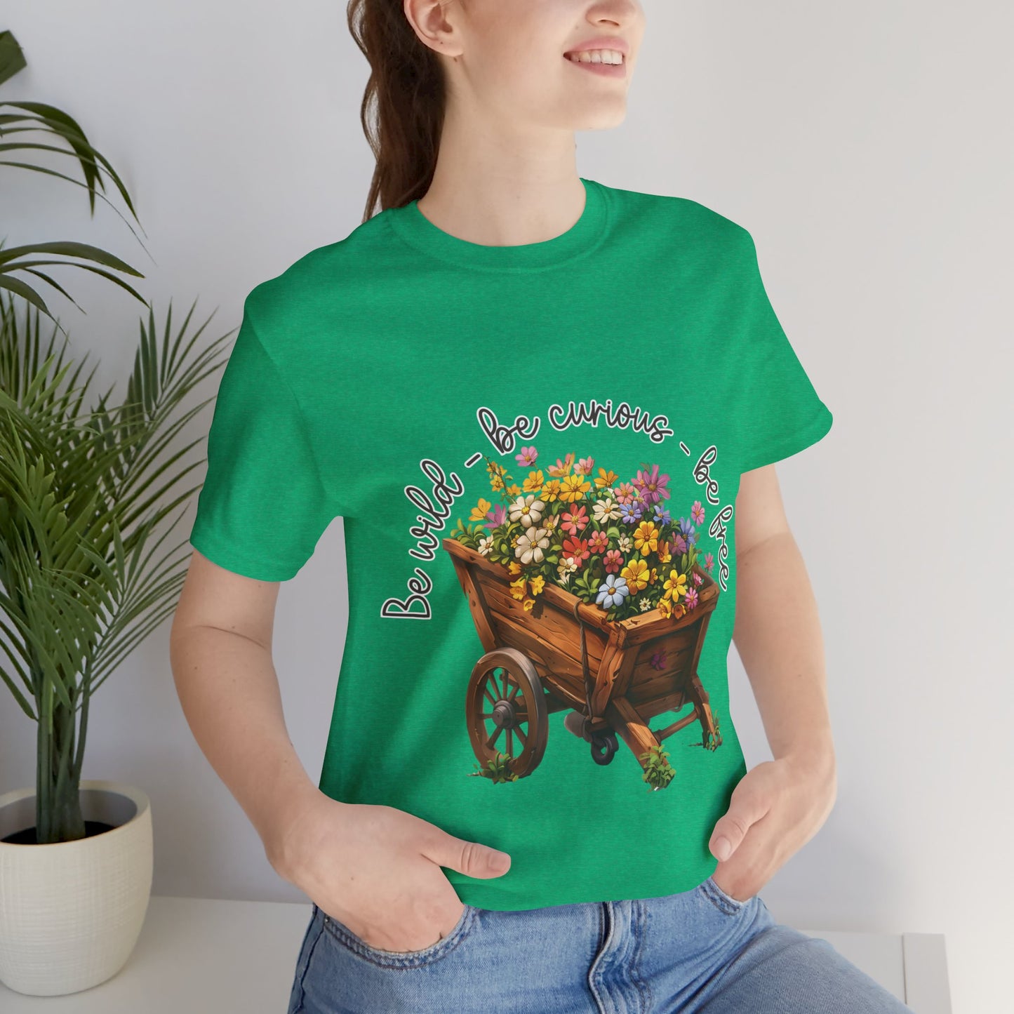 Colorful illustration of a wooden wheelbarrow filled with vibrant flowers, surrounded by the handwritten text 'Be wild - be curious - be free.' This design is intended for t-shirts in various colors, including Aqua, Athletic Heather, Black, Dark Grey Heather, Heather Kelly, Heather Red, Heather True Royal, Natural, Navy, Pink, White, Heather Aqua, Heather Ice Blue, Heather Mauve, and Charity Pink.