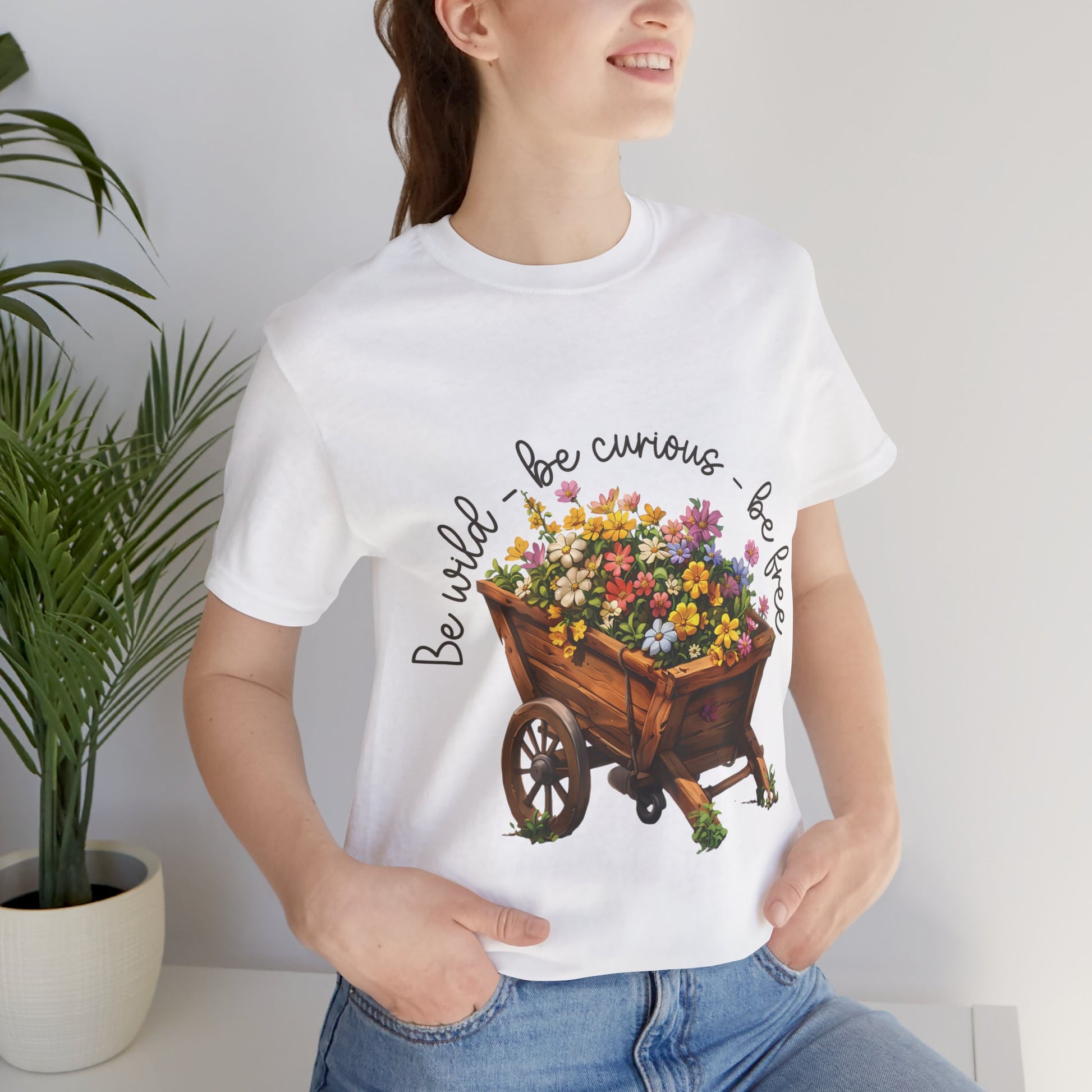 Colorful illustration of a wooden wheelbarrow filled with vibrant flowers, surrounded by the handwritten text 'Be wild - be curious - be free.' This design is intended for t-shirts in various colors, including Aqua, Athletic Heather, Black, Dark Grey Heather, Heather Kelly, Heather Red, Heather True Royal, Natural, Navy, Pink, White, Heather Aqua, Heather Ice Blue, Heather Mauve, and Charity Pink.