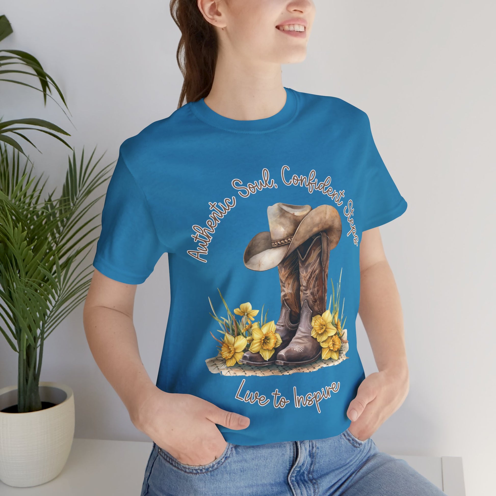 Brown leather cowboy boots with detailed stitching and a light brown hat on top, surrounded by green leaves and yellow flowers, with inspirational phrases ‘Authentic Soul,’ ‘Confident Steps,’ and ‘Live to Inspire’ written above and below on a white background colors tshirt Aqua Athletic Heather Baby Blue Black Dark Grey Heather Heather Kelly Heather Red Heather Team Purple Heather True Royal Navy Pink White Heather Aqua Heather Mauve Charity Pink.