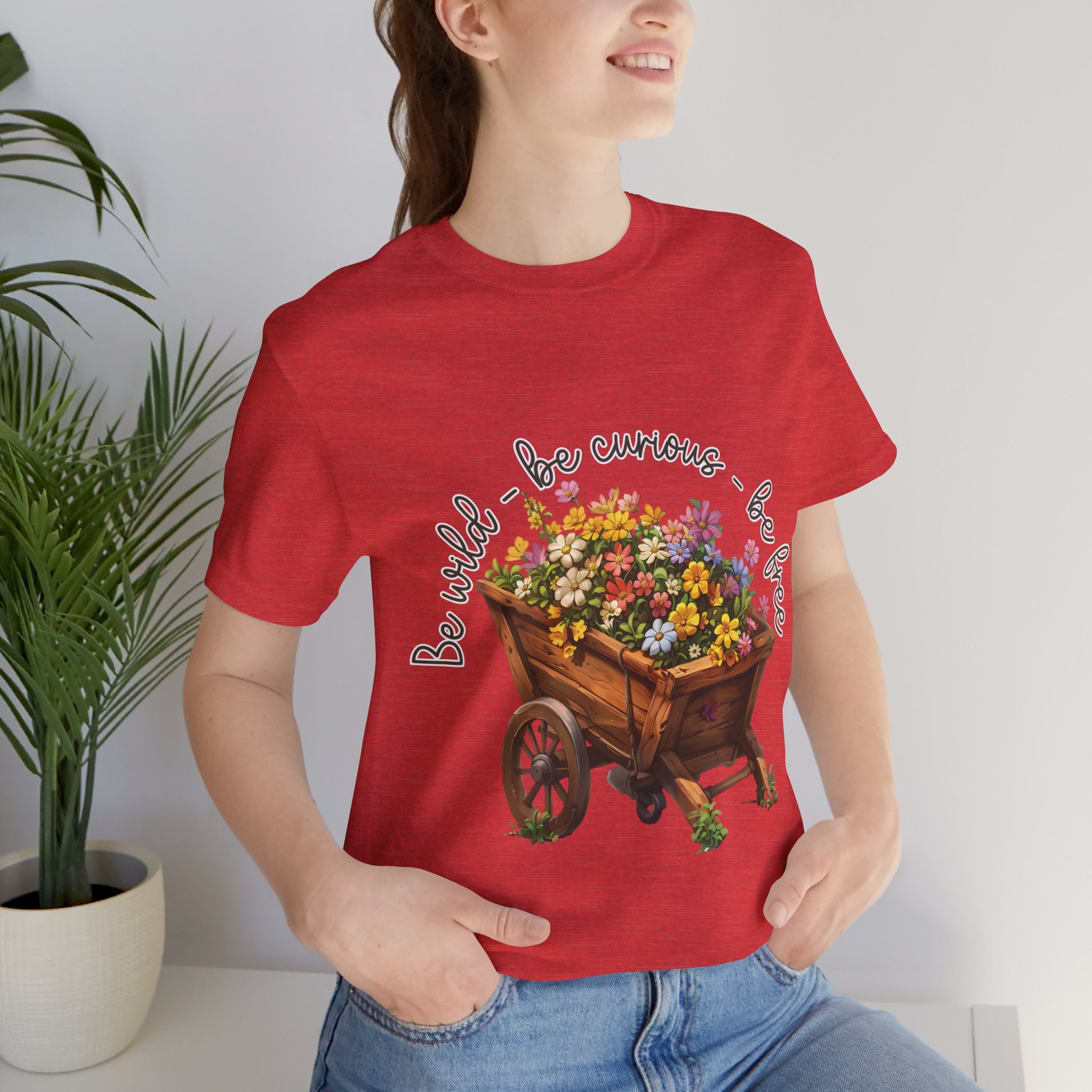 Colorful illustration of a wooden wheelbarrow filled with vibrant flowers, surrounded by the handwritten text 'Be wild - be curious - be free.' This design is intended for t-shirts in various colors, including Aqua, Athletic Heather, Black, Dark Grey Heather, Heather Kelly, Heather Red, Heather True Royal, Natural, Navy, Pink, White, Heather Aqua, Heather Ice Blue, Heather Mauve, and Charity Pink.