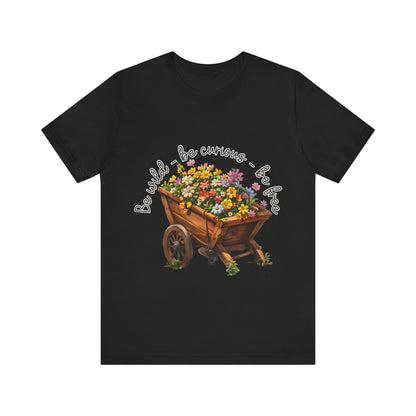 Colorful illustration of a wooden wheelbarrow filled with vibrant flowers, surrounded by the handwritten text 'Be wild - be curious - be free.' This design is intended for t-shirts in various colors, including Aqua, Athletic Heather, Black, Dark Grey Heather, Heather Kelly, Heather Red, Heather True Royal, Natural, Navy, Pink, White, Heather Aqua, Heather Ice Blue, Heather Mauve, and Charity Pink.