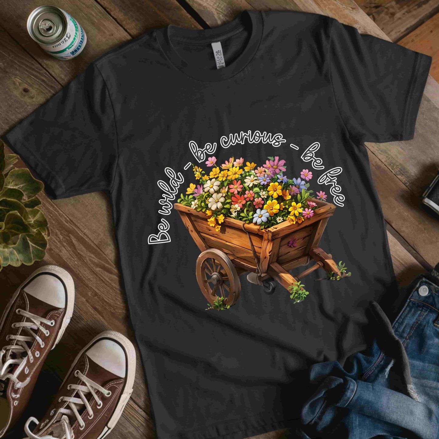 Colorful illustration of a wooden wheelbarrow filled with vibrant flowers, surrounded by the handwritten text 'Be wild - be curious - be free.' This design is intended for t-shirts in various colors, including Aqua, Athletic Heather, Black, Dark Grey Heather, Heather Kelly, Heather Red, Heather True Royal, Natural, Navy, Pink, White, Heather Aqua, Heather Ice Blue, Heather Mauve, and Charity Pink.